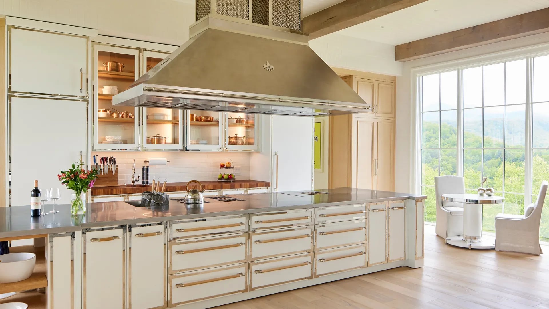 L'Atelier Paris white custom kitchen island with stainless steel countertop and a large professional ventilation hood featuring a fleur-de-lis emblem.